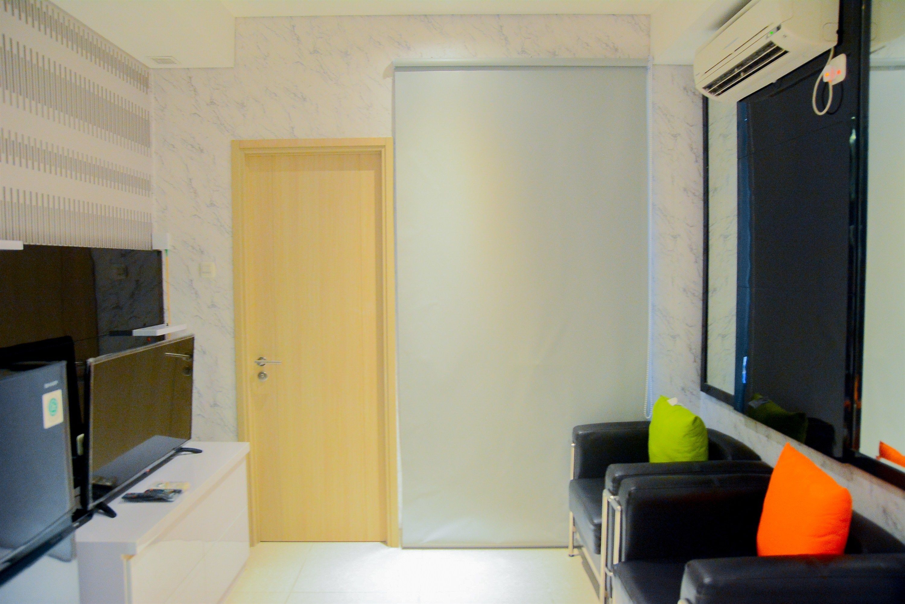 A hotel room with a closed door, a television on a stand, a sofa with a green and orange pillow, and an air conditioning unit mounted on the wall.