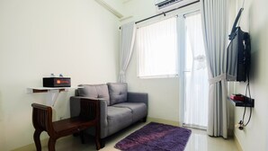 Ein Wohnzimmer mit grauem Sofa, einem Holzstuhl, einem violetten Teppich, einem an der Wand montierten Fernseher und einem Fenster mit Vorhängen.