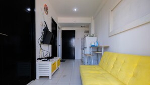 Ein modernes Wohnzimmer mit einem gelben Sofa, einer weißen Unterhaltungseinheit und einer Küchenzeile mit Kühlschrank und Wasserkocher.