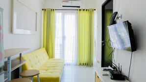 Ein Zimmer mit einem gelben Sofa, einem kleinen Tisch mit Hocker, einem Fernseher und einer Klimaanlage.