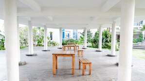 An open pavilion with wooden tables and benches, surrounded by white columns, set against a backdrop of greenery and a building.