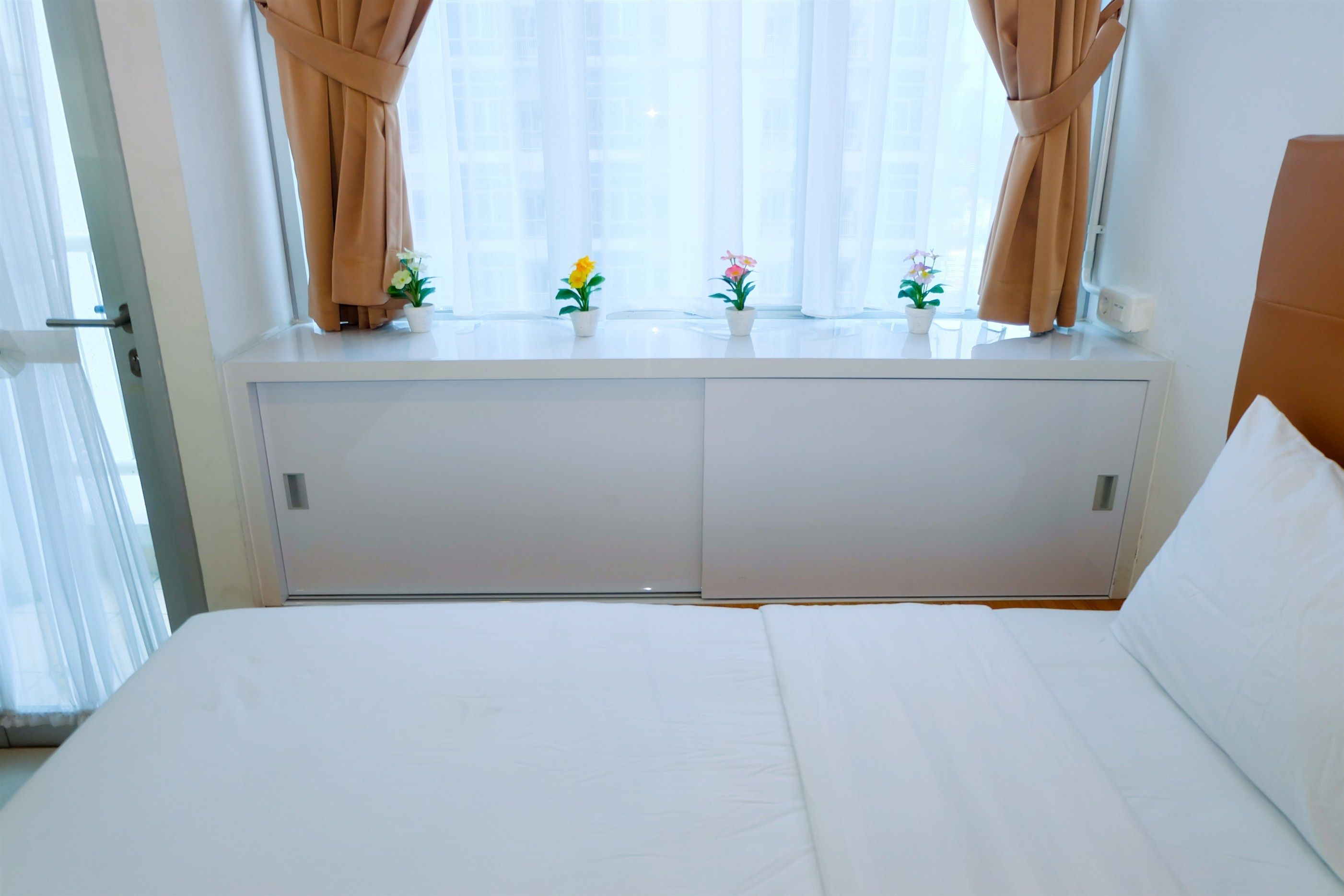 A hotel room with a bed, a window with curtains, and two small potted plants on the windowsill.