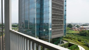 A high-rise building with a modern glass facade, a balcony with a metal railing, and a view of a landscaped area with greenery and construction equipment in the background. - Comfy Studio Ciputra International Apartment (Jakarta)