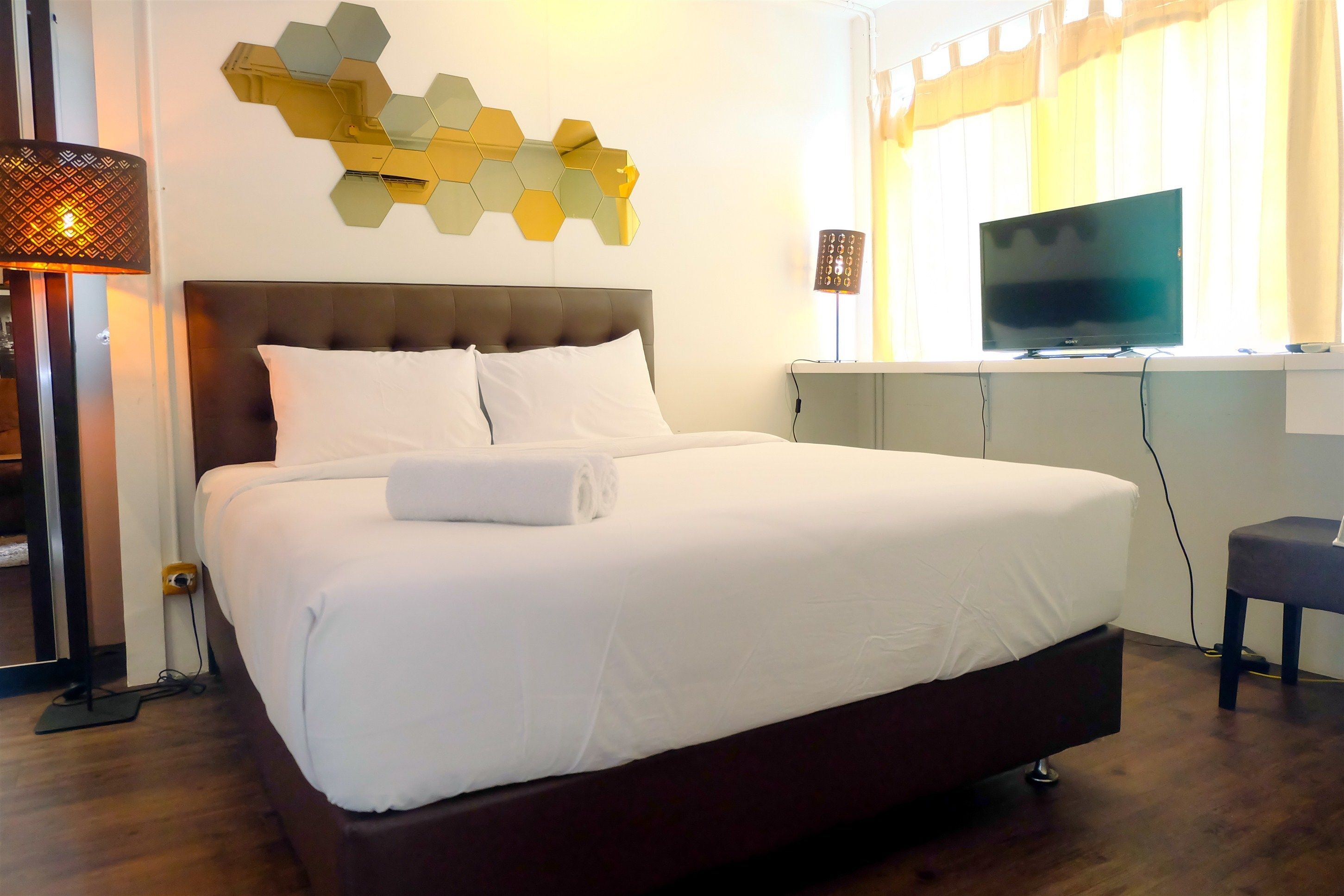 A hotel room with a large bed, a TV on a stand, a wall-mounted hexagonal mirror, and a lamp.