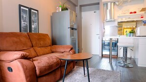 Ein modernes Wohnzimmer mit einem braunen Sofa, einem kleinen Holztisch und einem weißen Teppich. Im Hintergrund sind ein großer Kühlschrank sowie eine Küchenzeile mit Herd und Barhocker zu sehen.