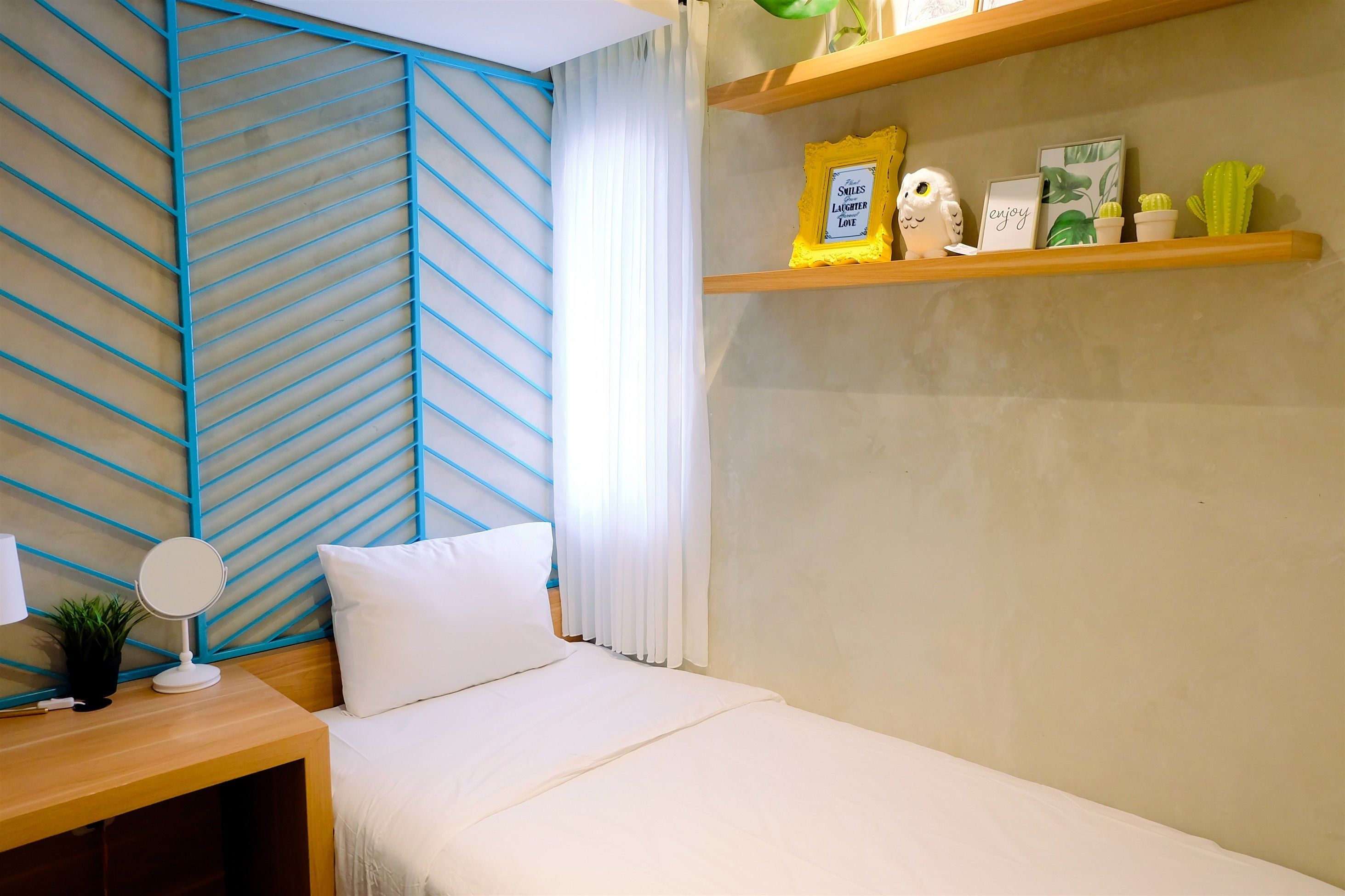 A neatly made bed with white bedding, a bedside table with a lamp, a small potted plant, and a wall with a blue geometric pattern.