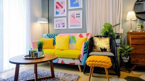 Un salón luminoso con un sofá amarillo, una mesa de centro redonda de madera y un sillón negro con un reposapiés amarillo.