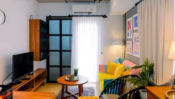 Una sala de estar moderna con un televisor de pantalla plana, muebles de madera, una mesa redonda de centro, cojines de colores y un ventanal con cortinas.