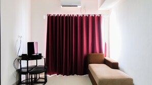 Een kleine, goed verlichte kamer met een bruine bank, een zwarte tv-standaard met een speaker, en een groot rood gordijn.