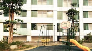 A playground with a swing set and slide, surrounded by a building with large windows and a paved area with a yellow line marking. - Fabulous Studio Grand Kamala Lagoon Apartment (Bekasi)