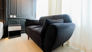 A modern living room with a dark-colored sofa, a small side table, and a white bed. - Fabulous Studio Grand Kamala Lagoon Apartment (Bekasi)