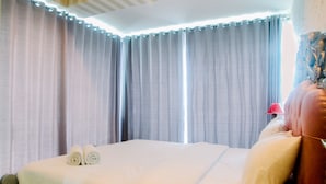 A hotel room with a neatly made bed, a red lamp on the nightstand, and a view of the outdoors through the curtains. - Luxury Furnished 2BR Grand Kamala Lagoon Apartment (Bekasi)