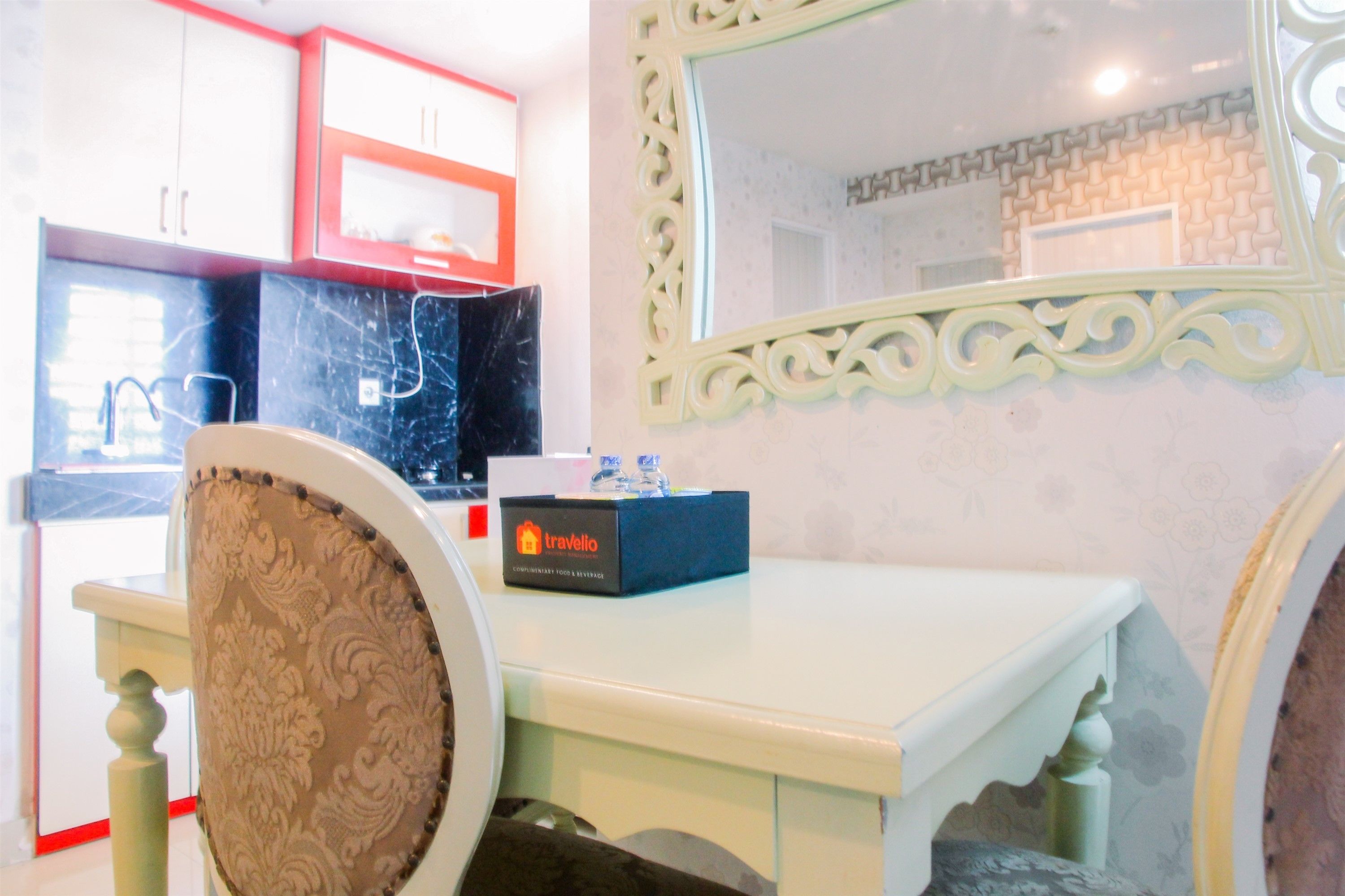 Un comedor con una mesa blanca, una silla con cojín estampado, una caja etiquetada 'travello' y armarios blancos y rojos al fondo.