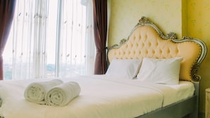 Un lit parfaitement fait, avec du linge de lit blanc et des serviettes pliées, une tête de lit capitonnée et une vue sur la ville à travers de fines rideaux.