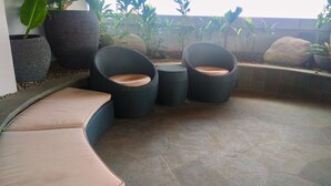 Un rincón de descanso con sillas de mimbre y un banco acolchado, rodeado de plantas en macetas y rocas.