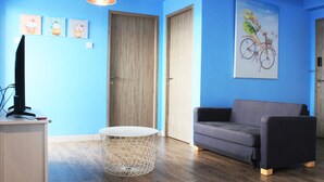 Une pièce avec un canapé gris, une table ronde blanche et un téléviseur posé sur un meuble. Il y a trois tableaux accrochés au mur, dont l’un représente un vélo.