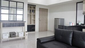 A modern living room with a black sofa, a wooden door, a built-in TV shelf, and a kitchen area with a refrigerator. - Cozy 2BR Apartment @ Newton Residence (Bandung)