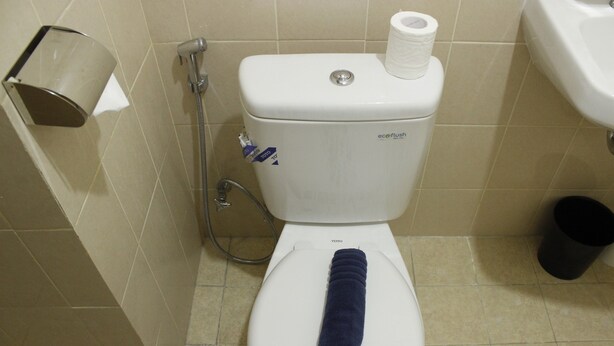 Un baño con inodoro blanco, una toalla azul sobre el asiento, dispensador de toallas de papel y un cubo de basura negro.