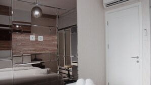 Una habitación de hotel con una cama, un espejo, un taburete pequeño y una puerta blanca.