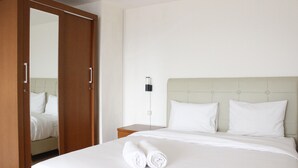 Una cama bien hecha con sábanas blancas y dos toallas enrolladas, un armario de madera y una mesita de noche con una lámpara.