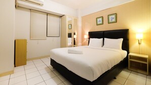 A hotel room with a large bed, a bedside table, a wall-mounted air conditioner, a window with blinds, and a wooden panel wall with framed pictures.