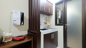 A compact kitchen with a water dispenser, a sink, and a microwave. There is a wooden shelf with kitchenware and a door with a frosted glass panel. - Minimalist Studio at Kebagusan City Apartment (Jakarta)
