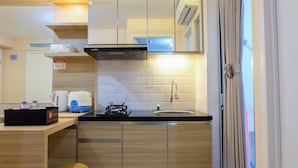 A compact kitchen with a gas stove, a sink, a microwave, and a coffee maker. There is a wooden countertop, a stainless steel range hood, and a mirror above the stove. A small stool is visible near the sink. - Clean 2BR Bassura City Apartment (Jakarta)