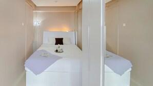 Una habitación pequeña con cama individual, colcha blanca, mesita de noche con lámpara y espejo en la pared.