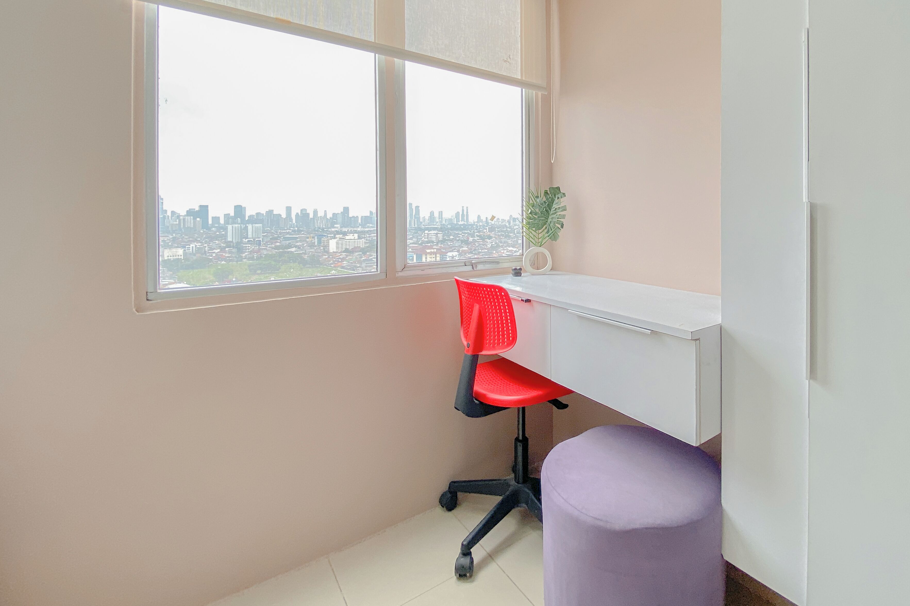 A room with a desk, a red office chair, a purple stool, and a window overlooking a cityscape.