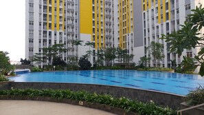 A swimming pool surrounded by greenery and a high-rise building with yellow accents. - Enjoy 2BR @ Springlake Apartment (Bekasi)