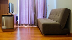 Ein Zimmer mit einem grauen Sofa, einem kleinen Fernseher auf einem Holzständer, einem GEA-Kühlschrank und einem Fenster mit violetten Vorhängen.
