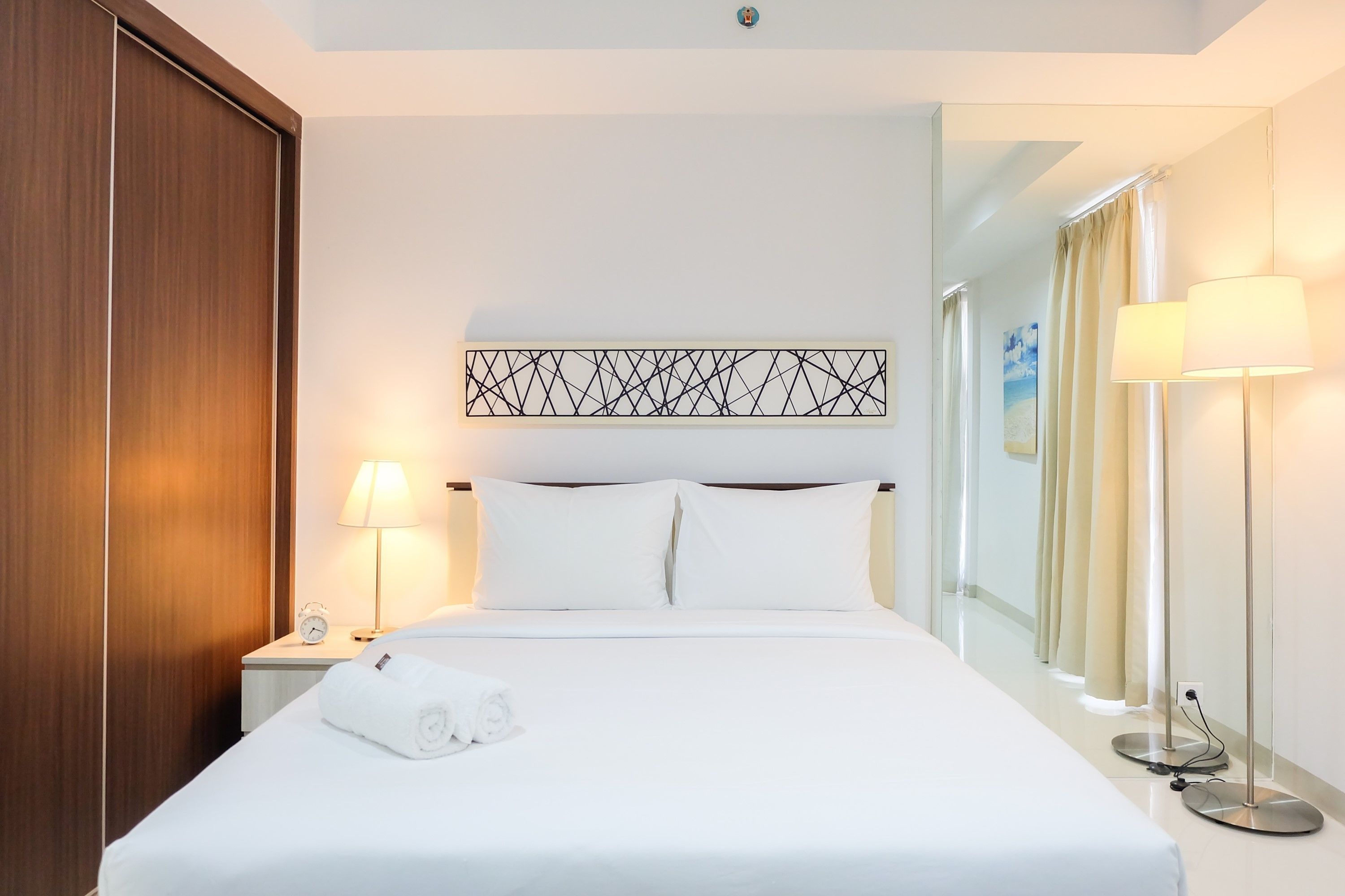 A hotel room with a large bed, a nightstand with a lamp, a wall clock, and a mirror.