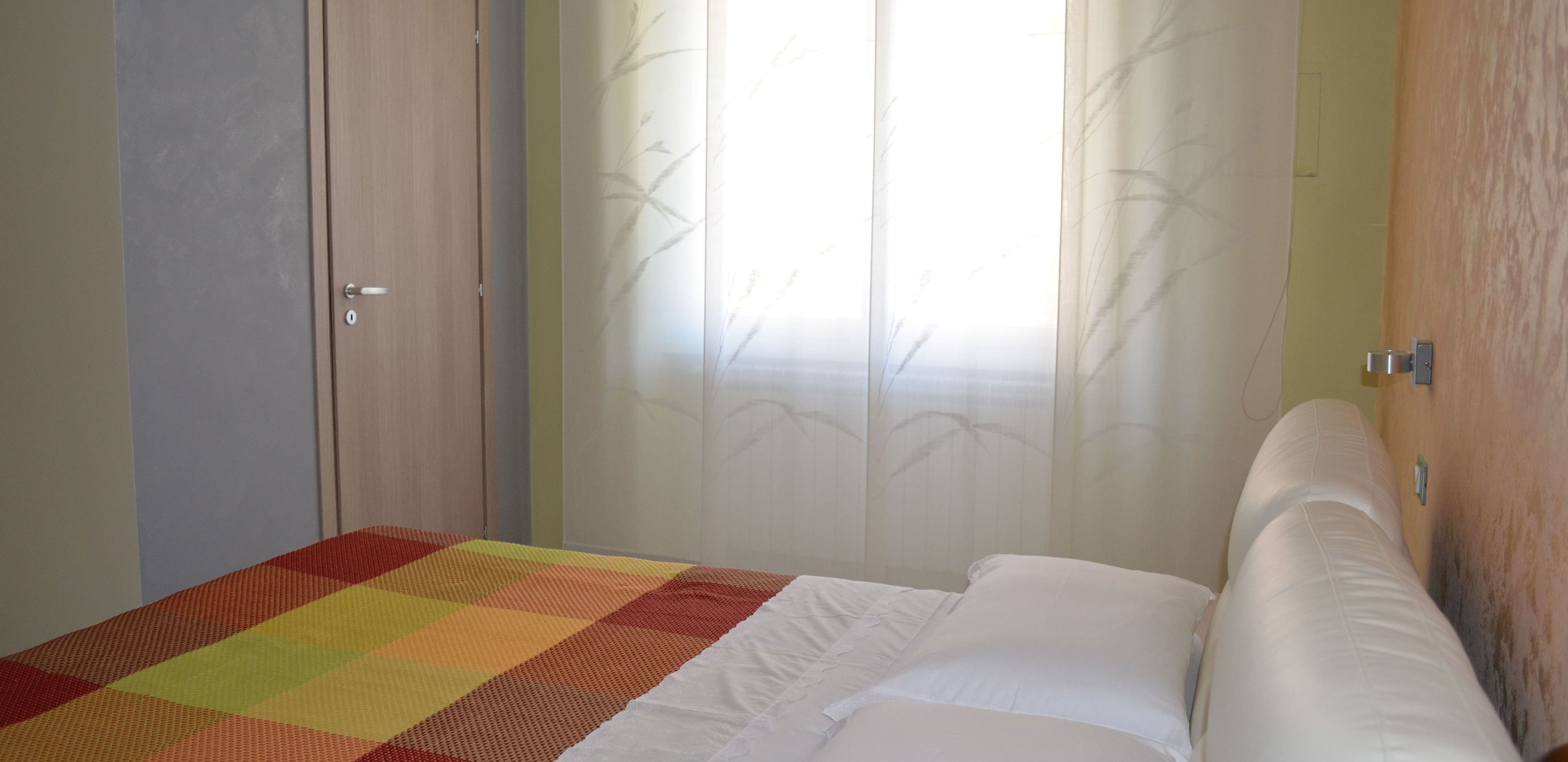 A bedroom with a bed, a colorful bedspread, a wooden wardrobe, and sheer curtains with a bamboo pattern.
