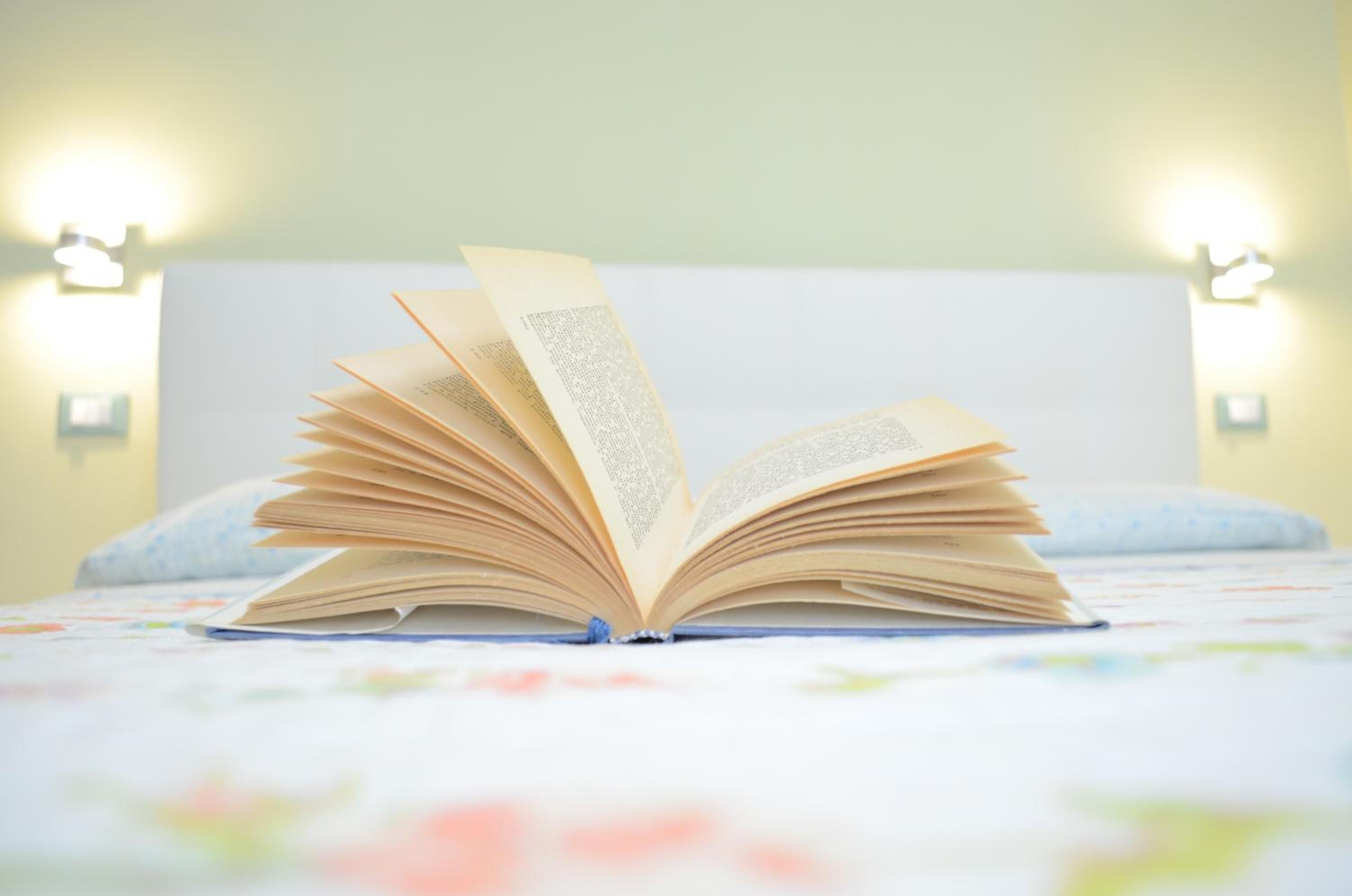 An open book on a bed with a white headboard, a floral-patterned bedspread, and two wall-mounted lamps.