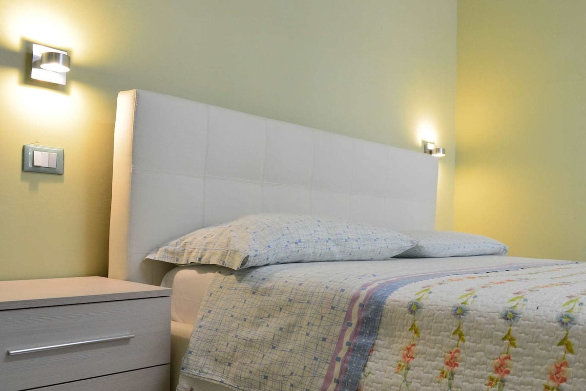 A neatly made bed with a floral quilt, a bedside table with a drawer, and two wall-mounted light fixtures.