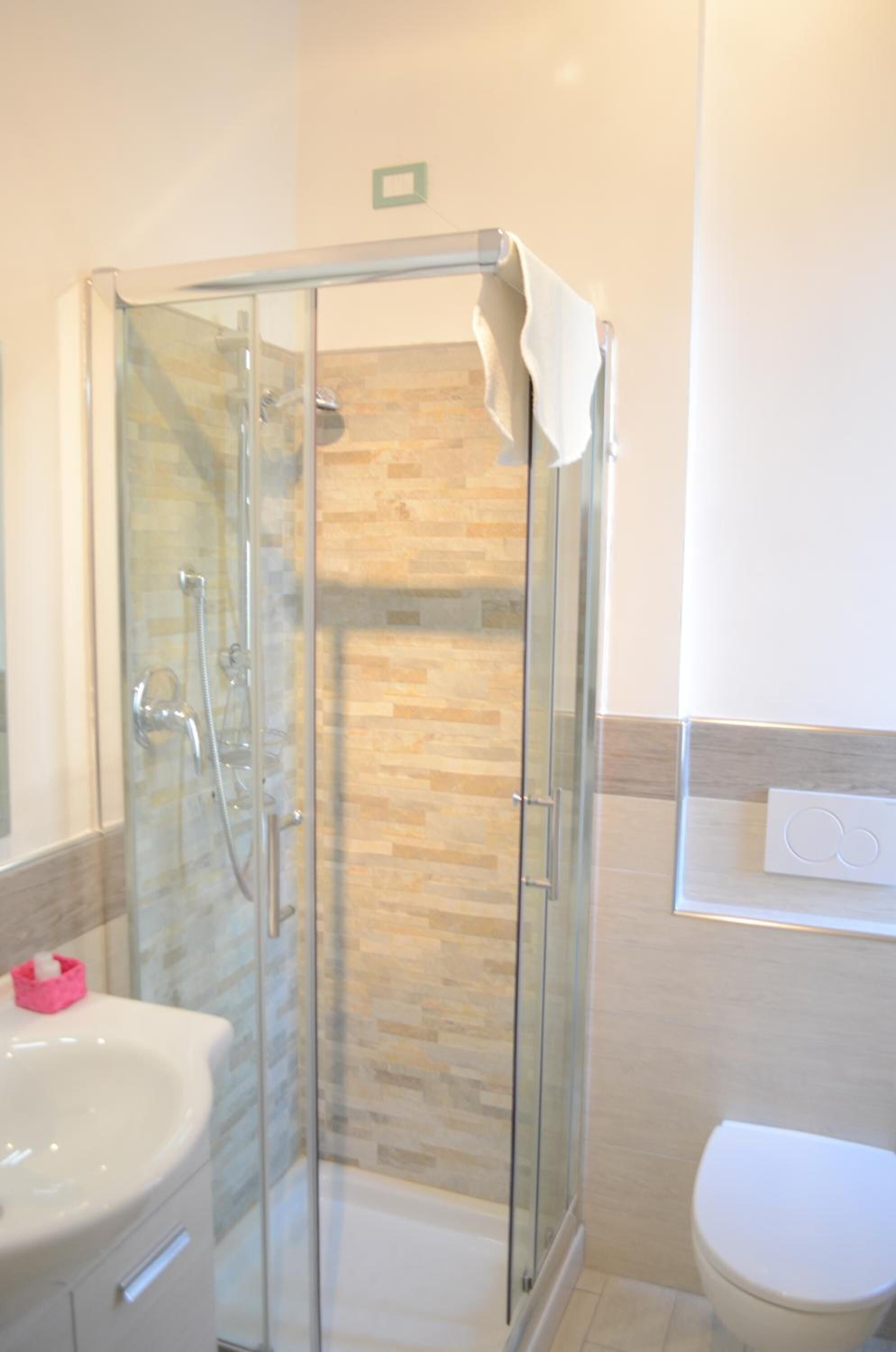 Ducha con cerramiento de vidrio, una toalla blanca colgada en la puerta, un lavamanos blanco y un inodoro en un baño con paredes de azulejos.