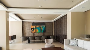 A modern living room with a sofa, coffee table, and chairs. There is a dining area with a table and chairs. The room has a large window and a decorative wall panel. - Elegant 1BR Branz BSD Apartment near AEON Mall (Pagedangan)