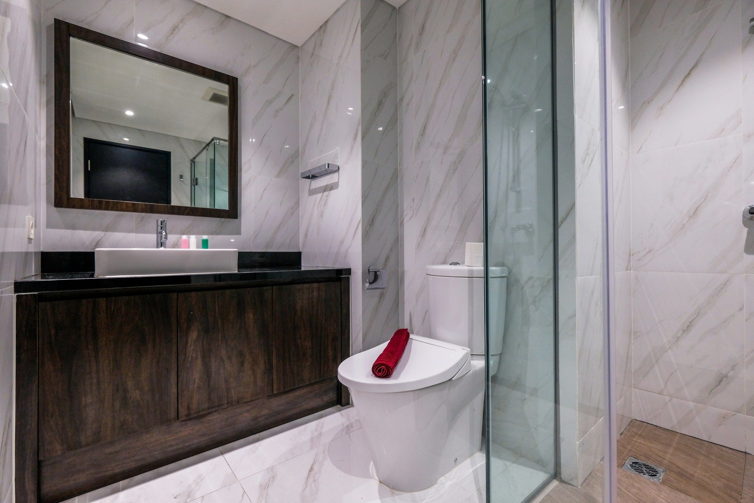 A modern bathroom with a marble vanity, a glass shower enclosure, and a toilet with a red towel on it.