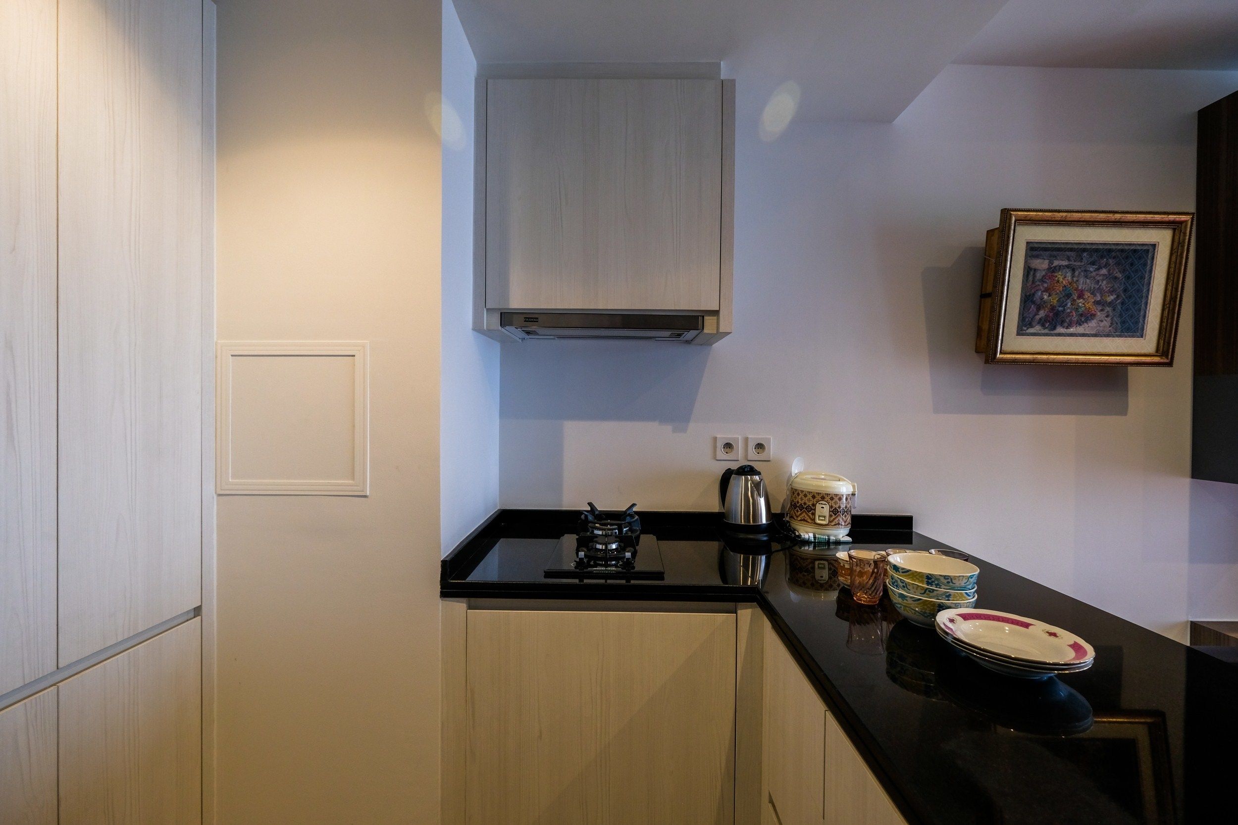 A compact kitchen with a black countertop, a stainless steel kettle, and a coffee maker. There are two wall-mounted light switches and a framed picture on the wall.