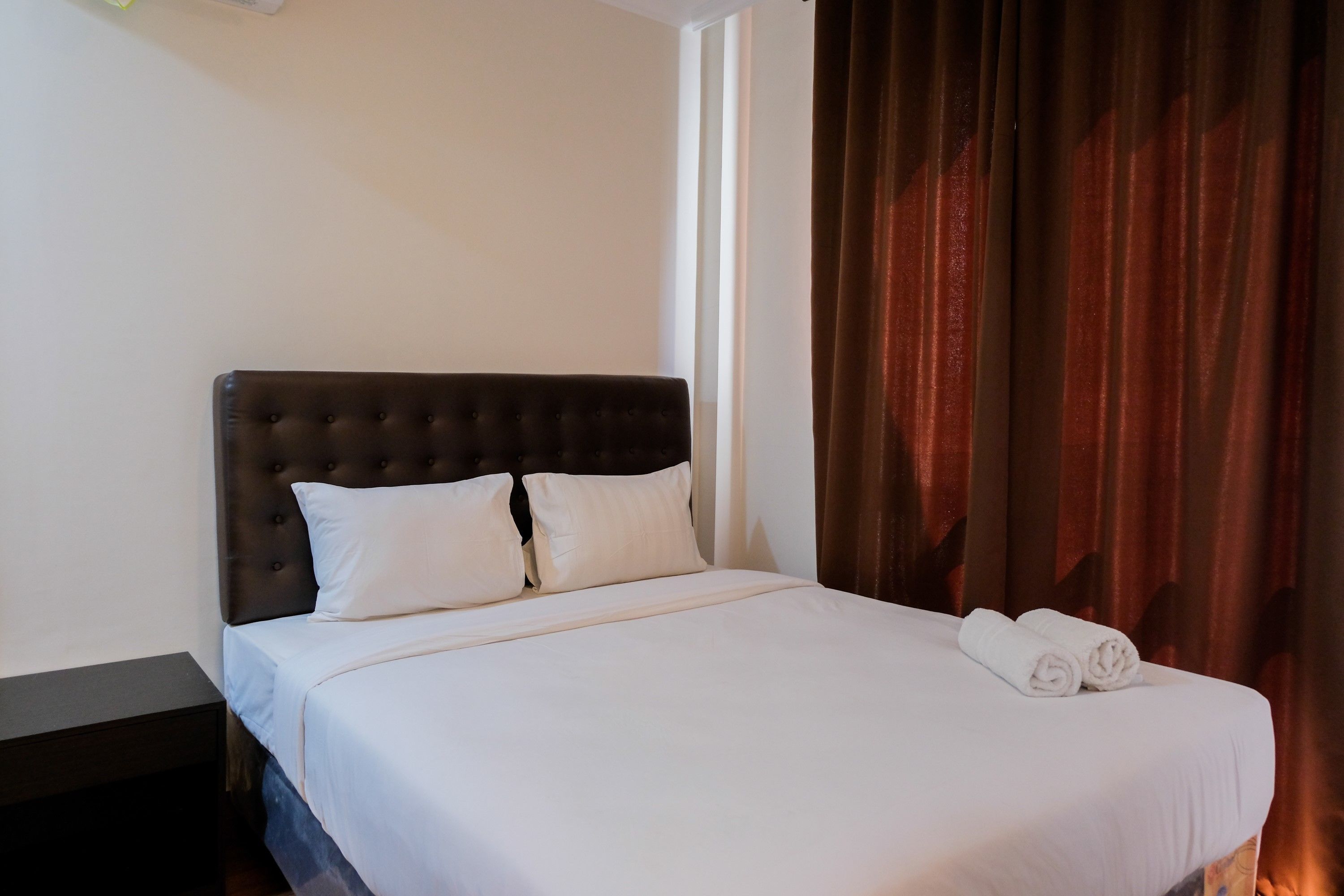 A neatly made bed with white linens and two pillows, a dark headboard, and a pair of rolled towels on the foot of the bed.