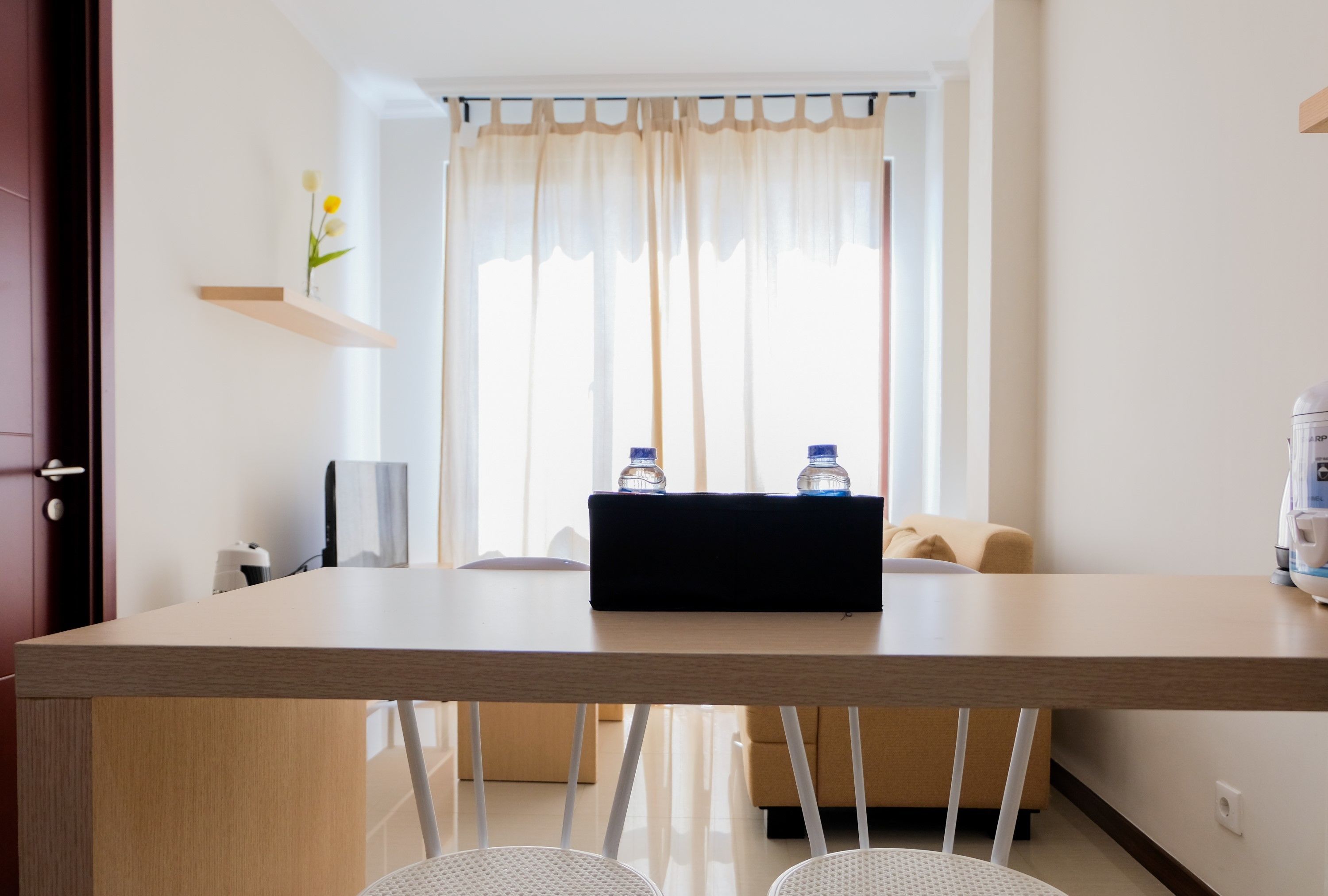 Een moderne hotelkamer met een houten eettafel, twee stoelen, een bank, een klein plankje met een bloem en een raam met gordijnen.