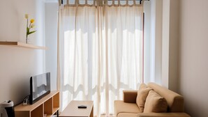 Ein modernes Wohnzimmer mit einem beigefarbenen Sofa, einem Flachbildfernseher auf einem Holzboden, einem Couchtisch und transparenten Vorhängen.
