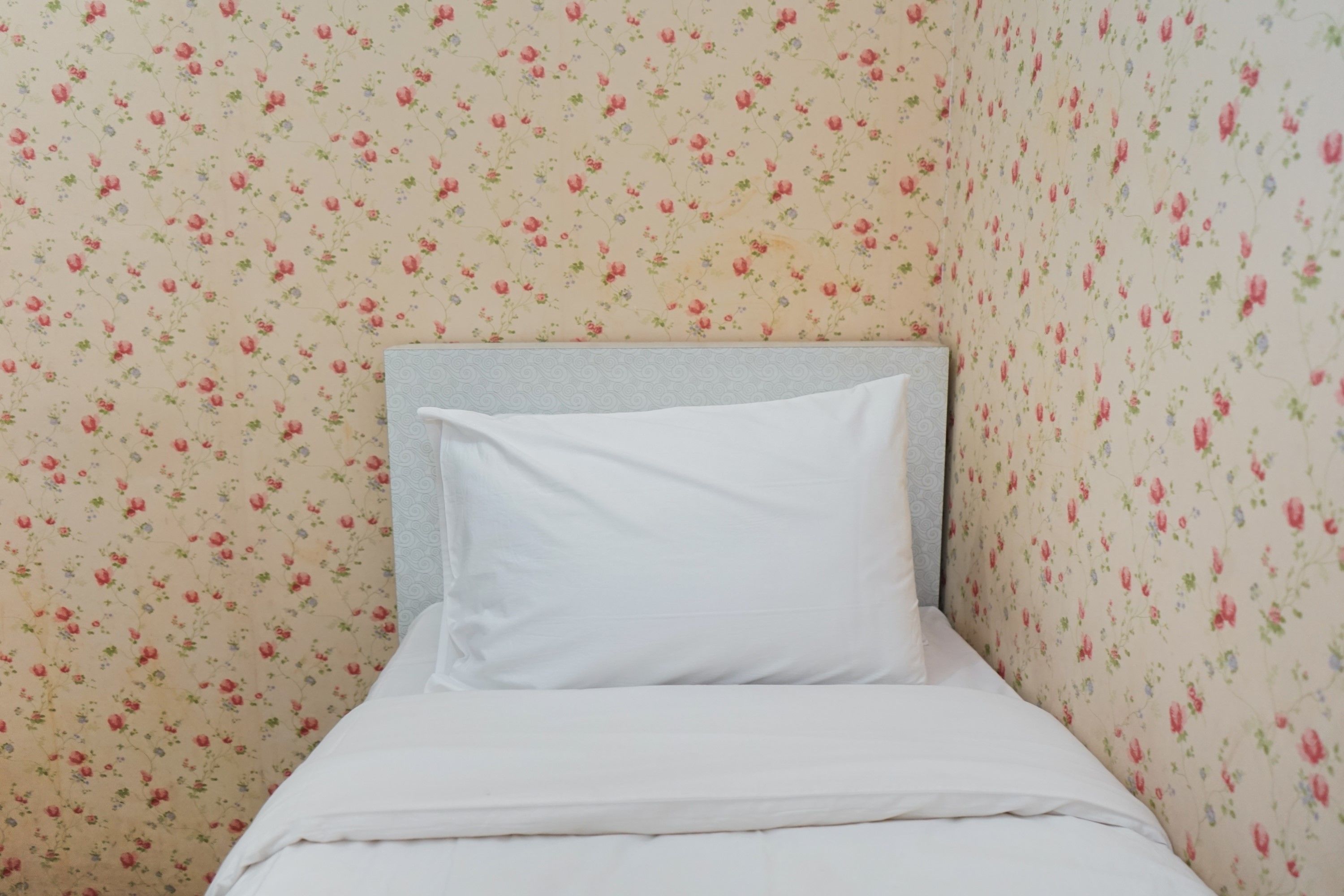 A single bed with a white pillow and a grey headboard in a room with floral wallpaper.