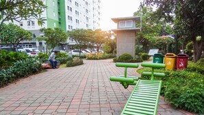 A paved walkway with benches and trash bins, surrounded by greenery and a building in the background. - Comfy and Clean 2BR Green Pramuka Apartment (Jakarta)
