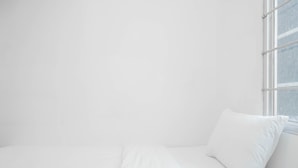 A white bed with a single white pillow in a room with white walls and a window.