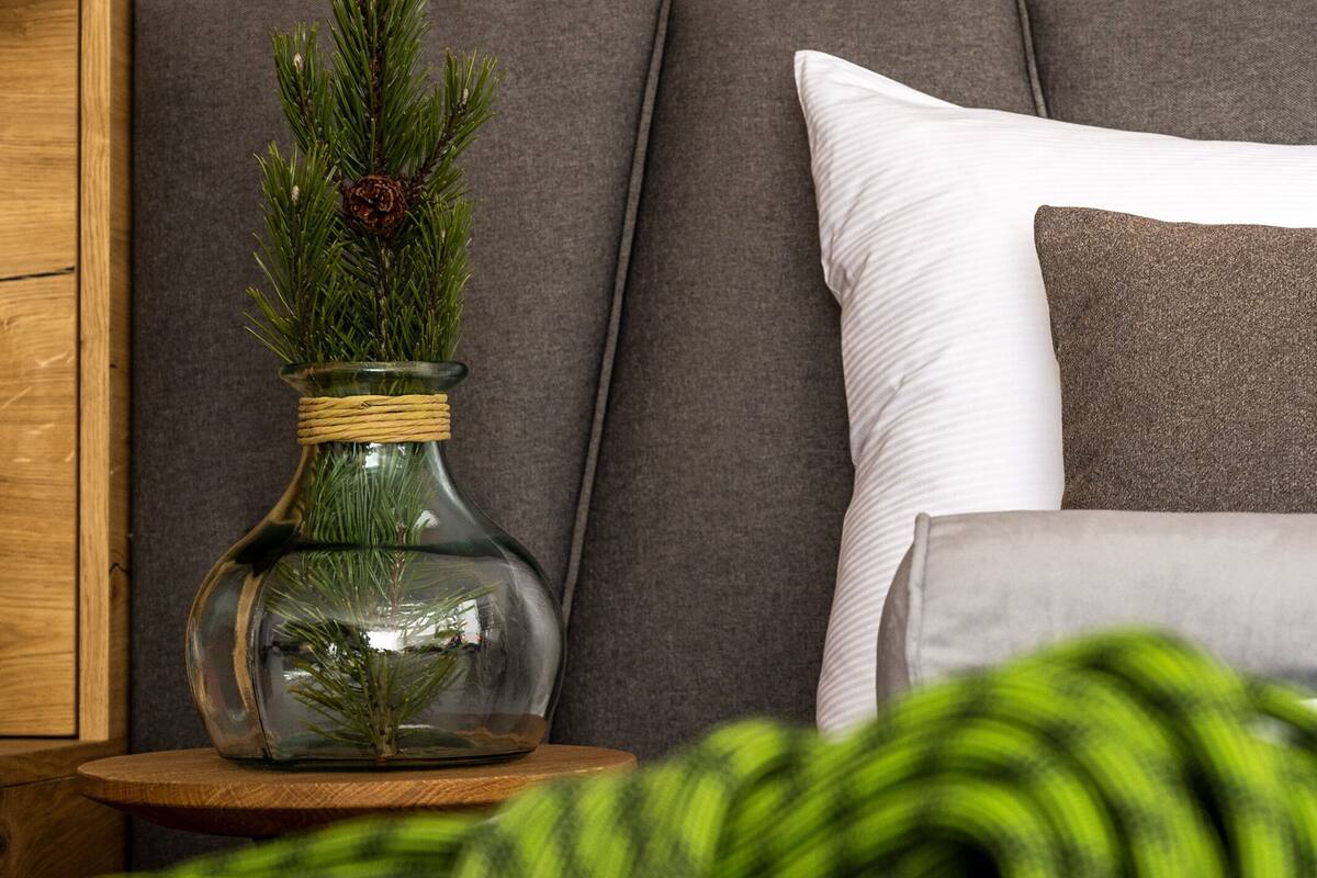 Un dormitorio moderno con cabecera gris, un florero de vidrio con ramas de pino, una mesita de noche de madera y una manta verde.