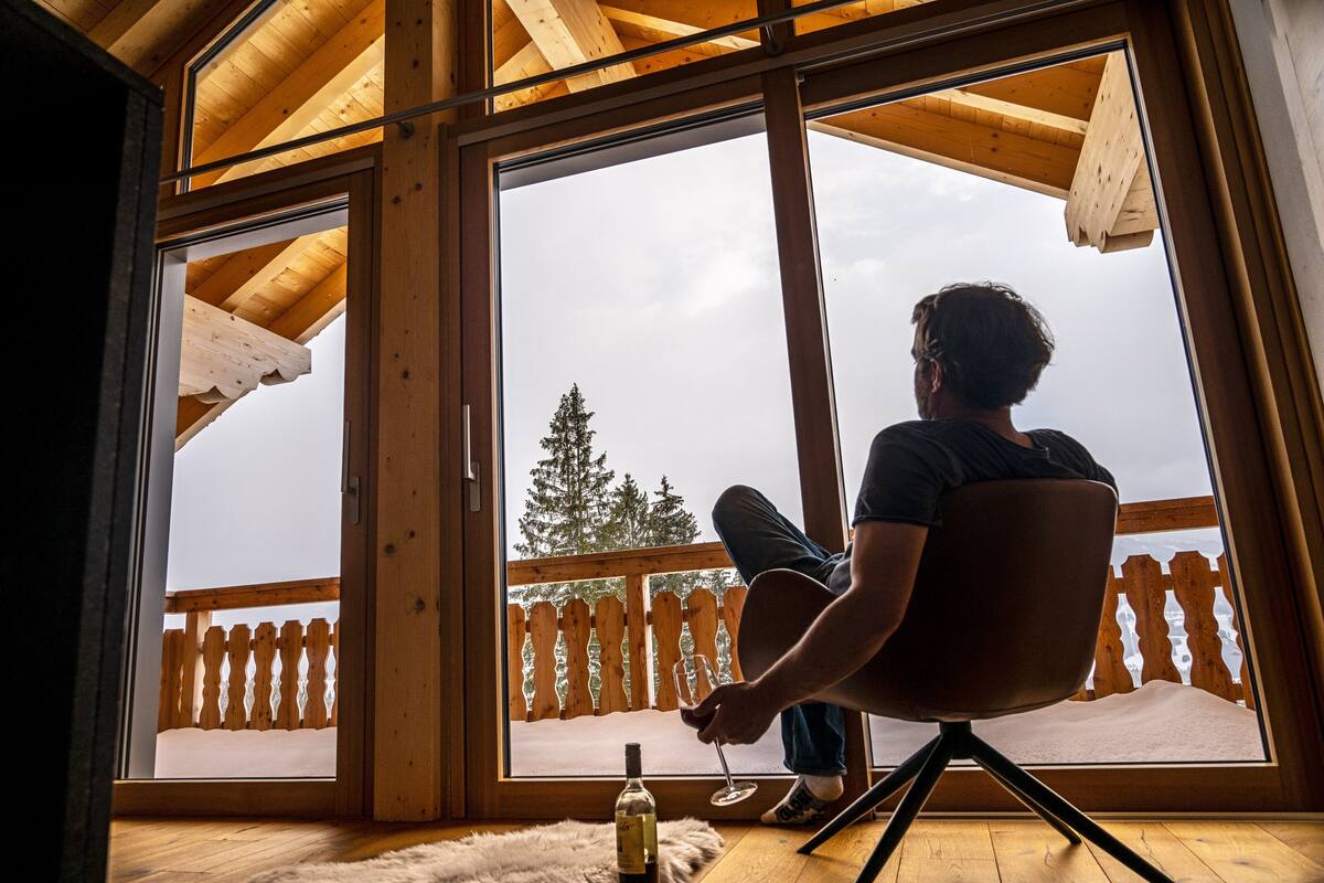 Una persona sentada en una silla frente a un ventanal grande con vistas a una terraza de madera y un árbol.