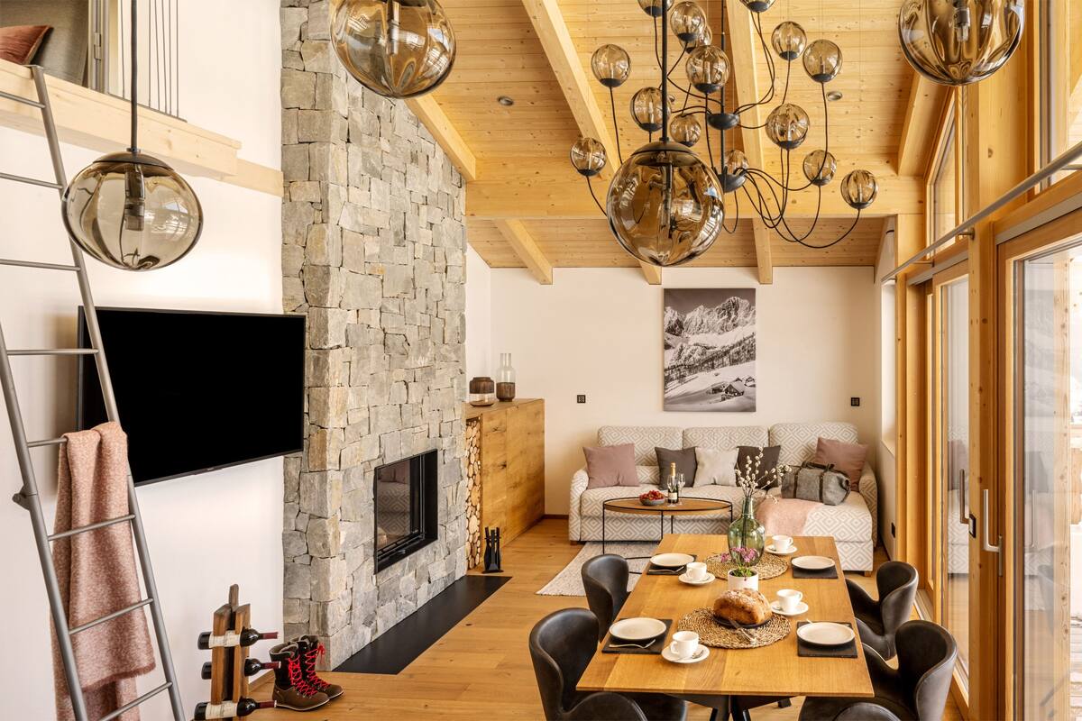 Una sala de estar moderna con chimenea de piedra, vigas de madera en el techo y un comedor con mesa y sillas de madera.