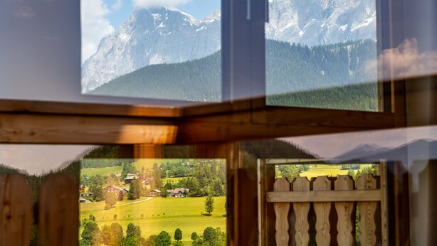 A view through a window with a wooden frame, displaying a mountainous landscape with snow-capped peaks and a valley with houses and greenery. - Alpine Residence Dachsteinperle (Ramsau am Dachstein)