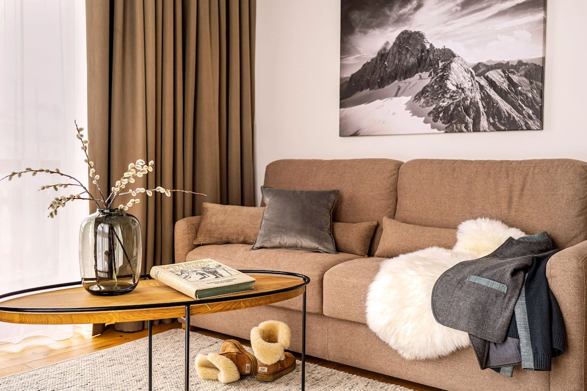 Una sala de estar acogedora con un sofá marrón, una mesa de centro de cristal, un libro, un jarrón con ramas y un cuadro de un paisaje montañoso en la pared.